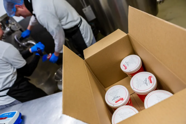 Ice cream containers in cardboard box, workers in background.